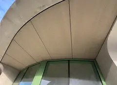 Gebogen plafond van gevelplaten bij een modern gebouw, met een deel van groene ramen zichtbaar.
