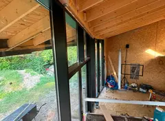 Binnenaanzicht van houten plafond en grote ramen met tuin op de achtergrond, bouwmateriaal op de vloer.