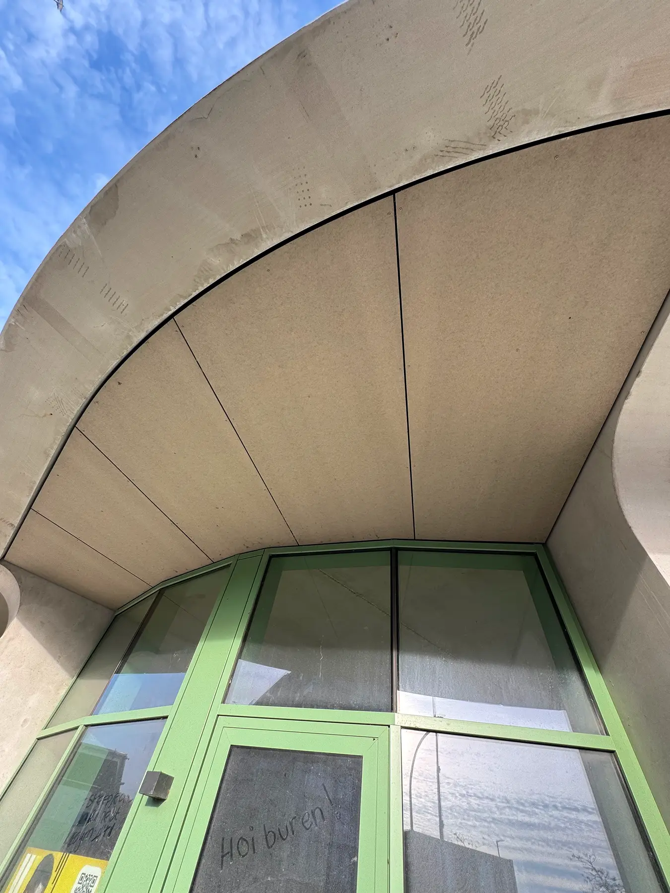 Gebogen plafond van gevelplaten bij een modern gebouw, met een deel van groene ramen zichtbaar.