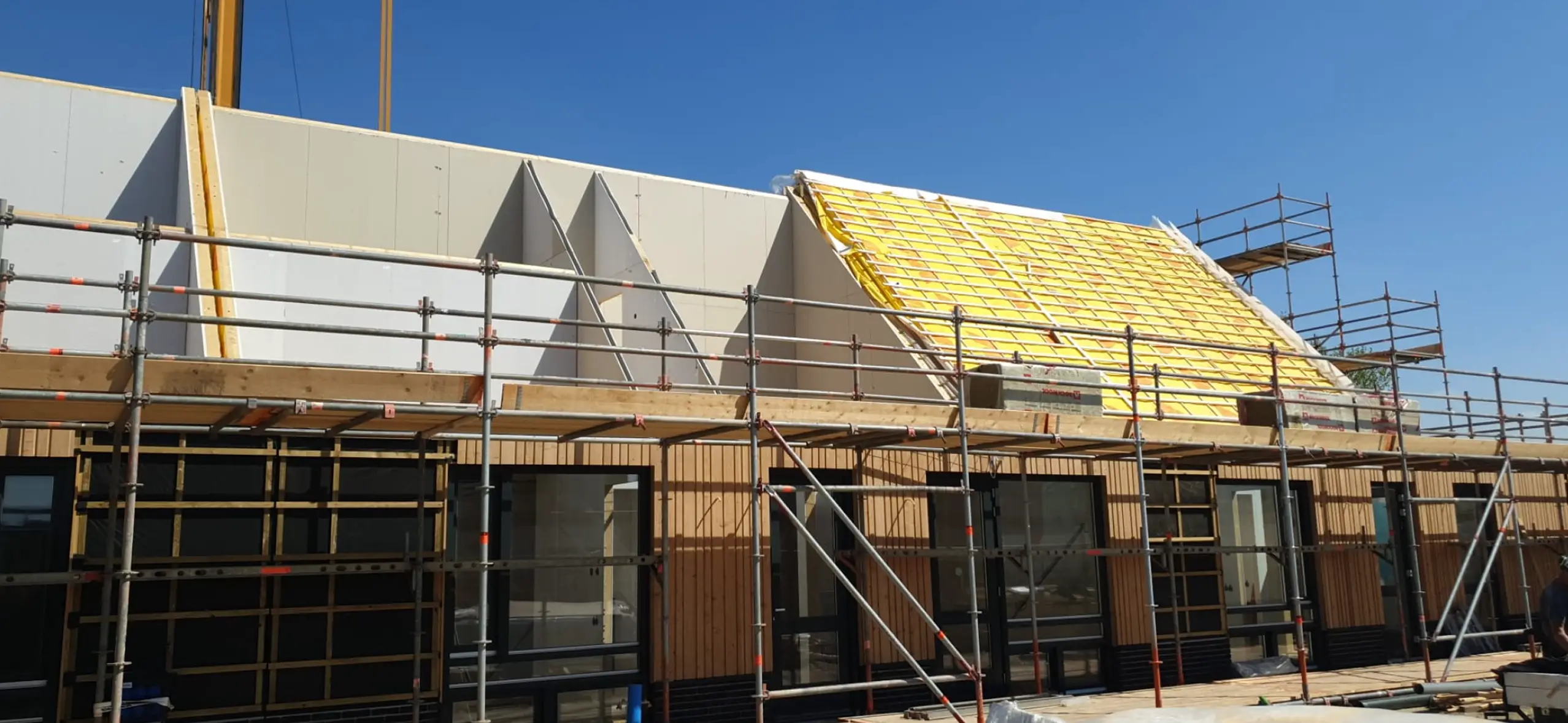 Gebouw in aanbouw met steigerwerk, houten gevel en montage van een dak met gele isolatie.