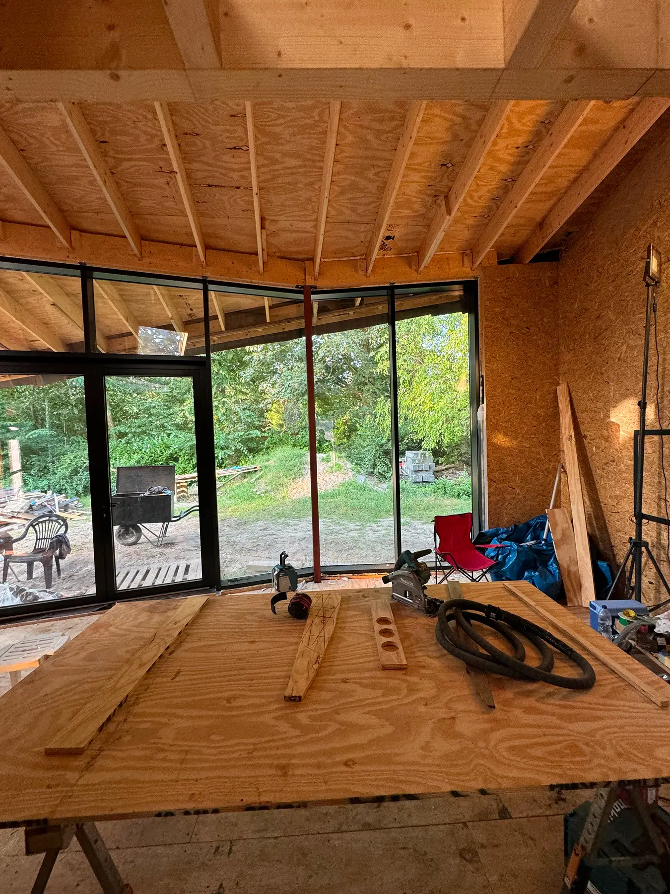 Interieur van een houten constructie met werktafel, gereedschap en grote ramen met uitzicht op het groen.