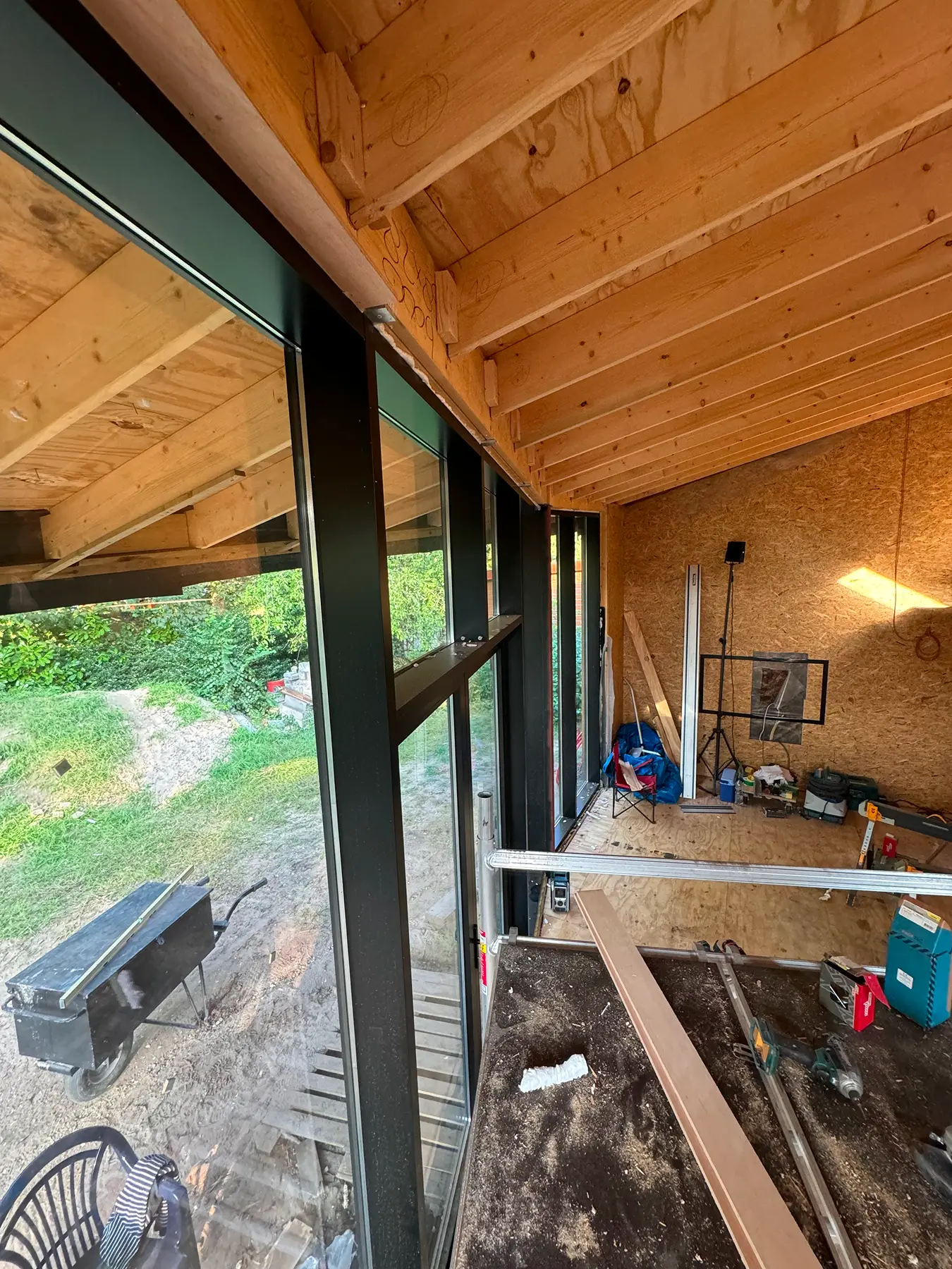 Binnenaanzicht van houten plafond en grote ramen met tuin op de achtergrond, bouwmateriaal op de vloer.