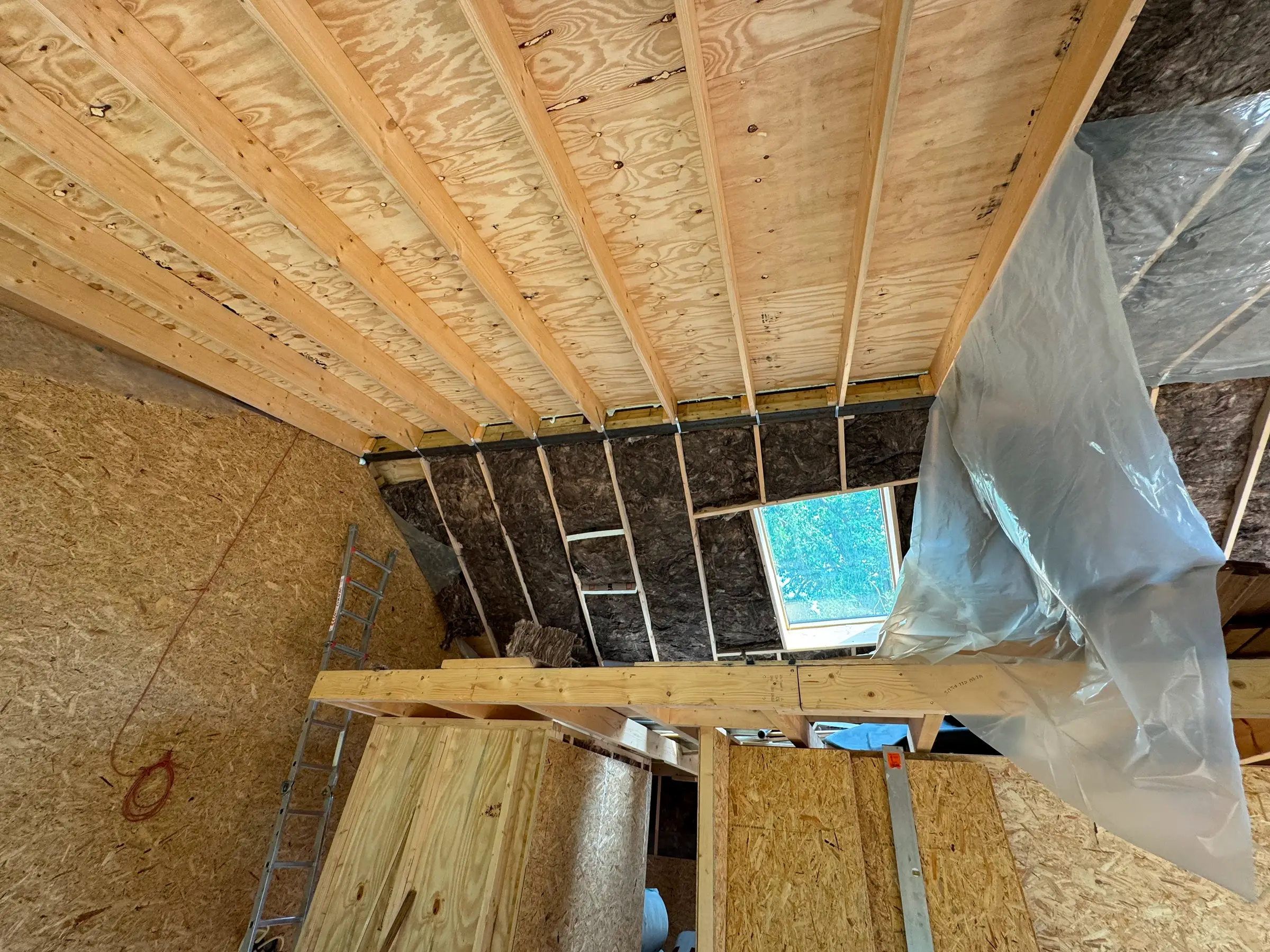 Schuin houten plafond met isolatie en gedeeltelijk aangebrachte dampremmende folie, ladder en dakraam zichtbaar.