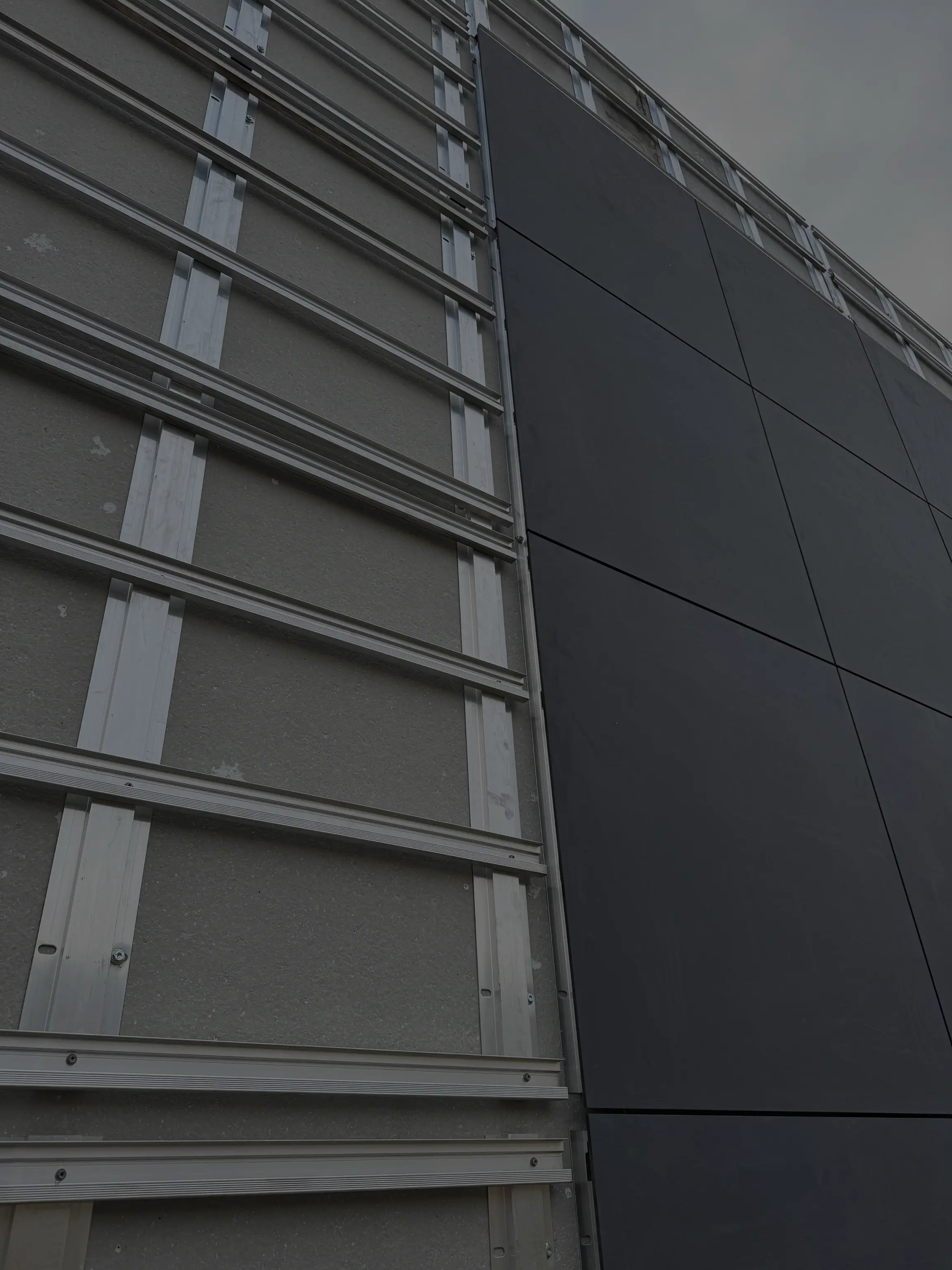 Een fragment van een gebouwgevel tijdens de montage van een geventileerde gevel; aan de linkerkant zijn aluminium draagprofielen bevestigd aan de isolatielaag, en aan de rechterkant zijn donkere gevelpanelen geplaatst die het afgewerkte oppervlak vormen.